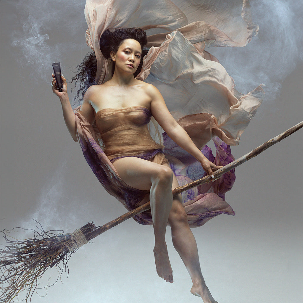 A surreal photograph of a woman floating on a broomstick with flowing sheer fabric, holding the Flying Ointment salve.