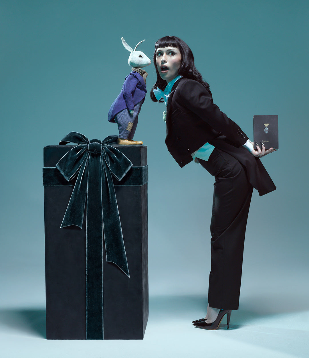 A woman in a black tuxedo leans over a large black gift box with a velvet bow, while a small rabbit figurine in a purple suit stands on top of the box. She holds a Rhubarb Thief Discovery Set.