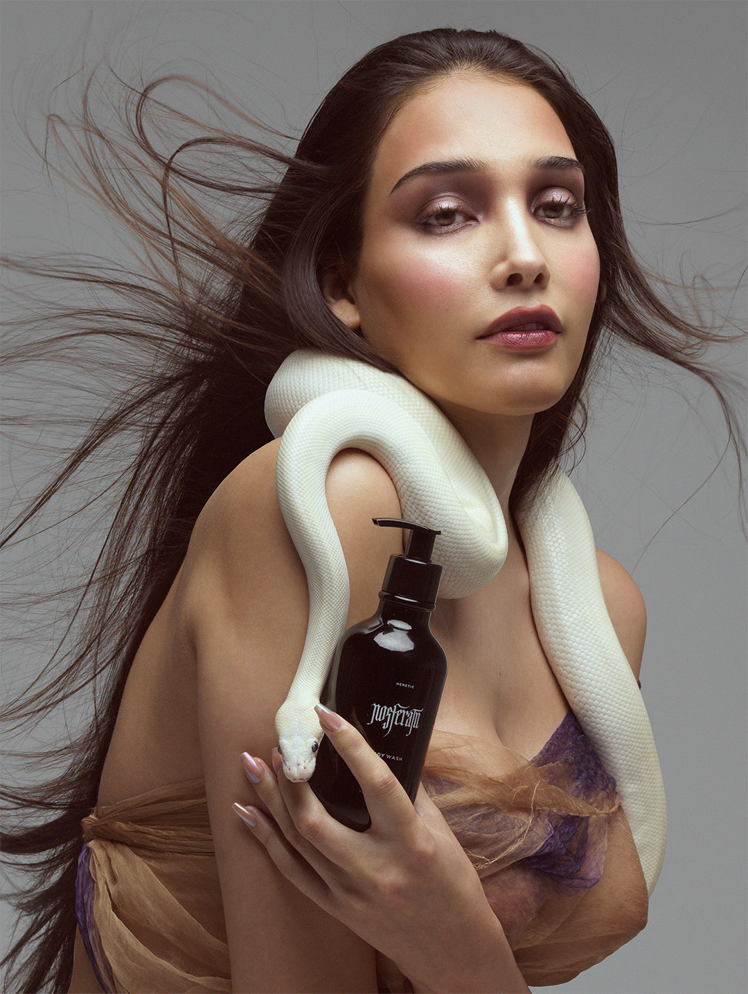 A woman with long dark hair poses with a white snake draped over her shoulders and holds a black bottle.